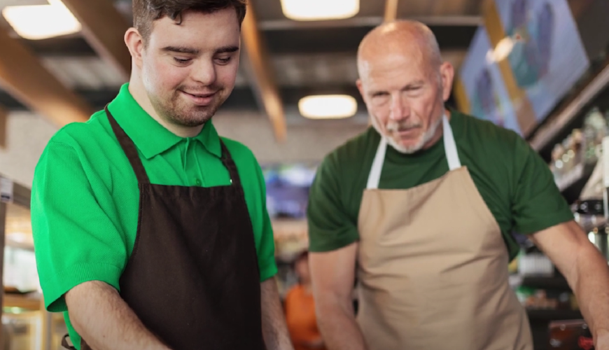 Image of 2 men wearing aprons in a cafe. Both men are wearing green shirts and one is older than the other. The setting is a cafe and the two men are depicted as cafe staff. 