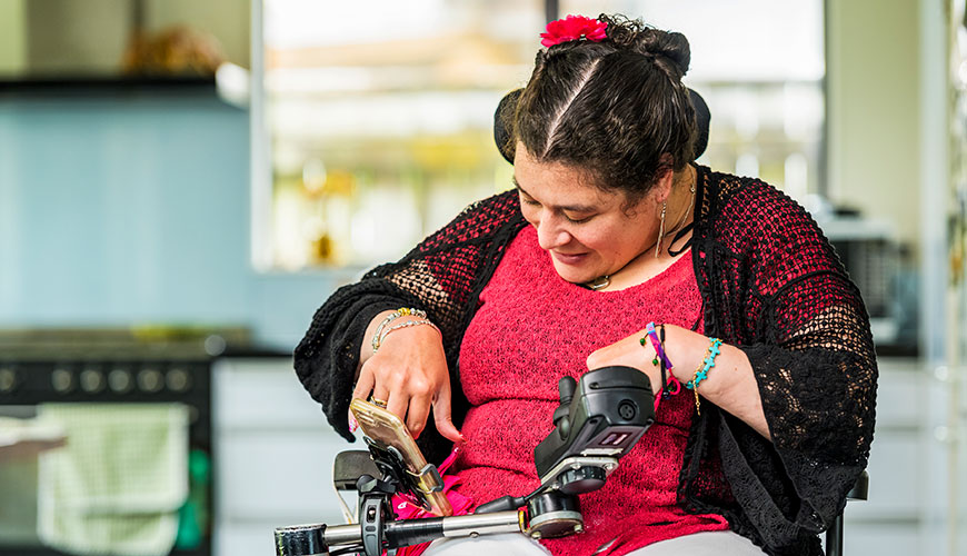 A woman dressed in a red top and black shawl is using her mobile phone. The phone is mounted to her electric wheelchair.
