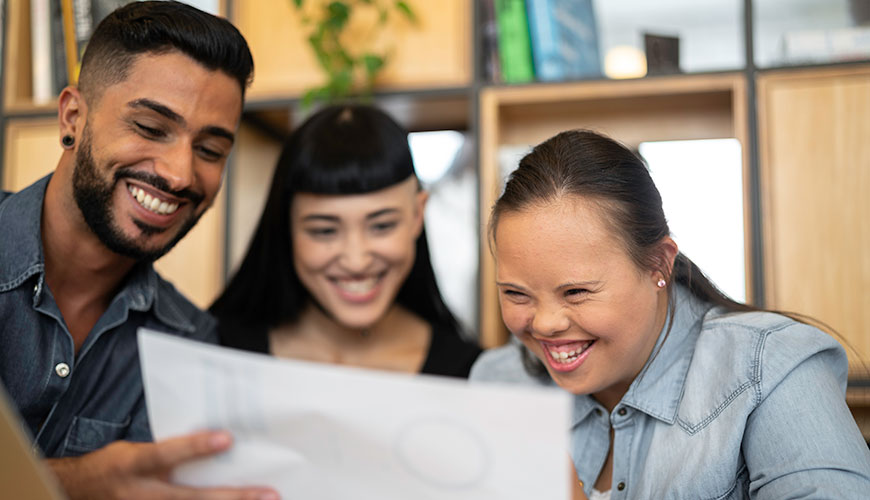 Three people smiling and looking at a work plan together. From left to right: There is a man with dark skin and an earring, a woman with long black hair and another woman also with long dark hair, which is pulled back, who appears to have Down Syndrome.