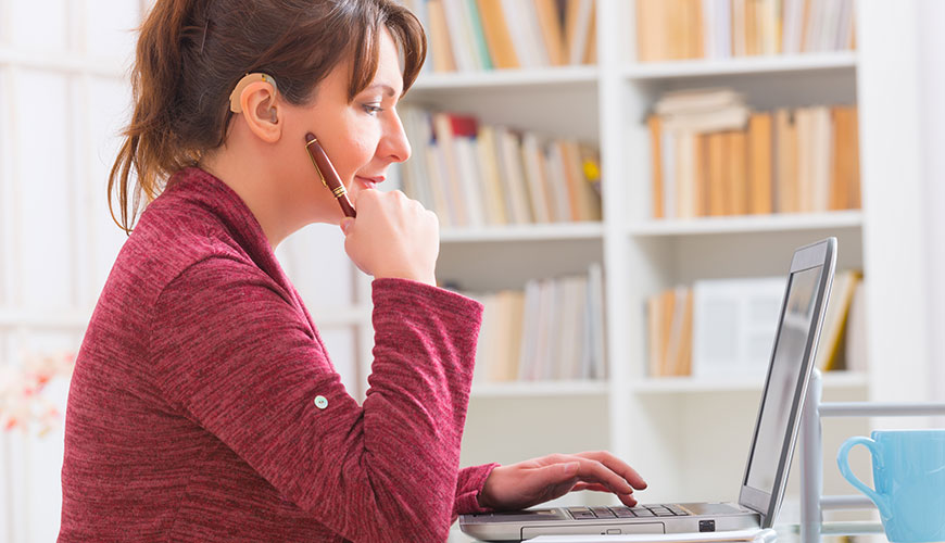 A woman sitting at a desk who is working on her computer. She is dressed in red and has a hearing device.