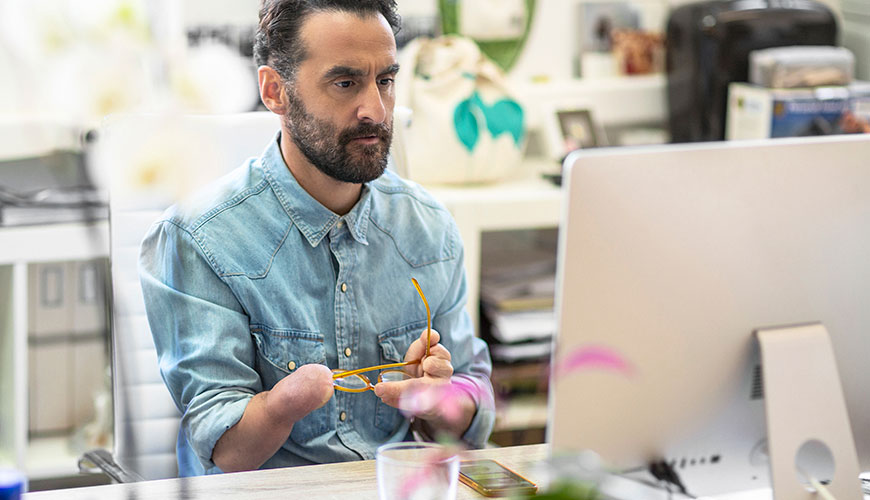 A man sitting at a desktop computer. He holds his glasses between his arms as he has no hand.