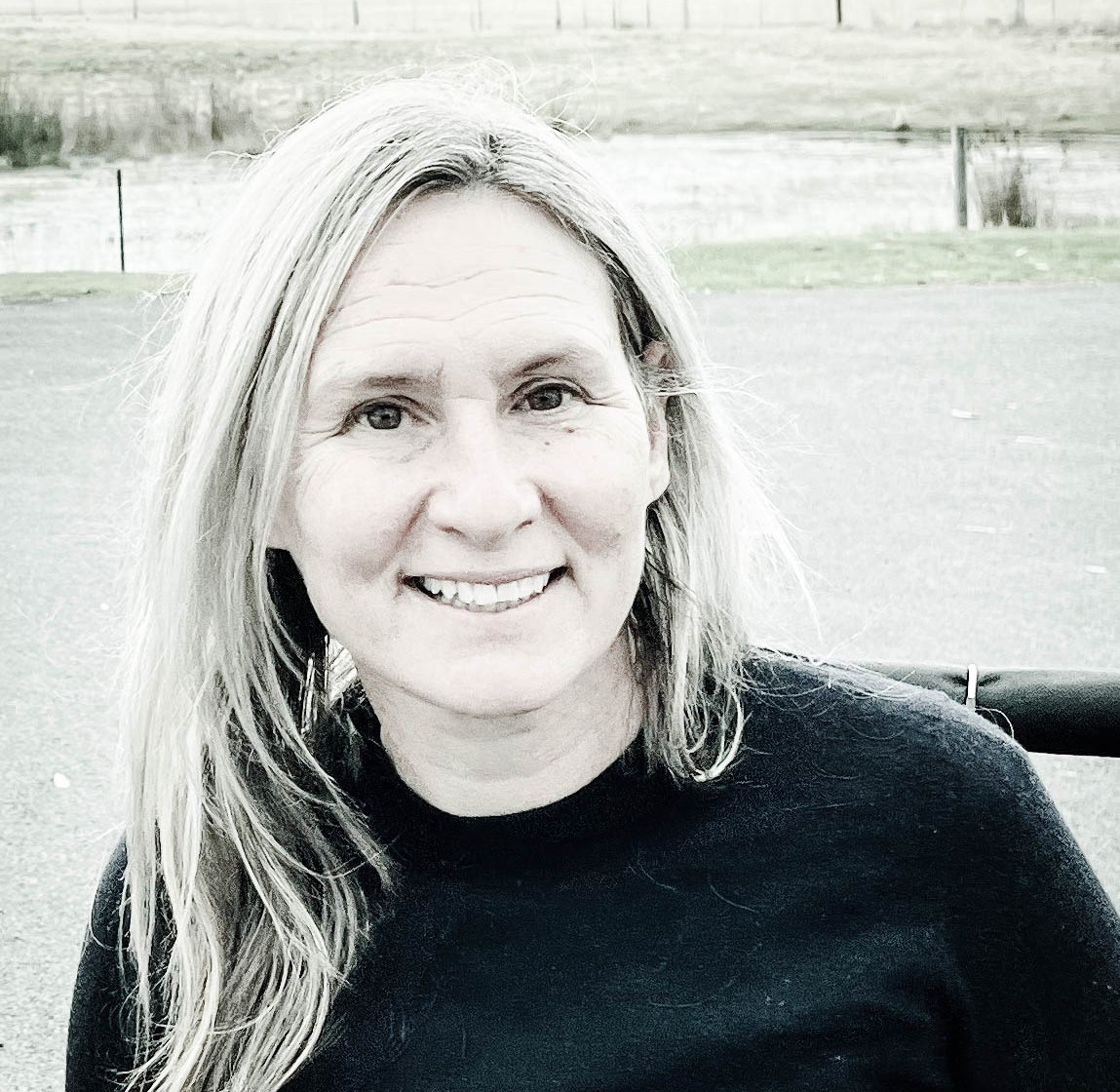 Image of Lisa Stafford wearing a black jumper and smiling. Lisa has long blond hair. The background is farm land. 