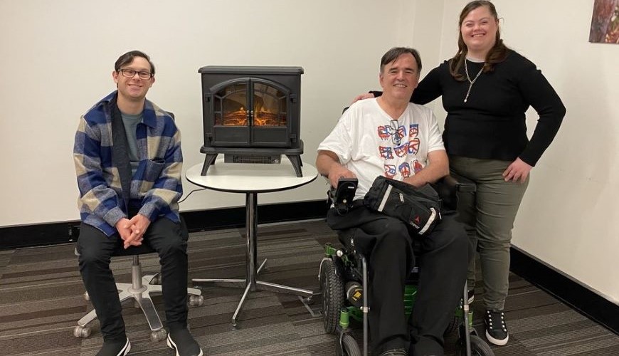 image of three people and a fireplace. They are smiling at the camera. One of the people is sitting in a wheelchair.