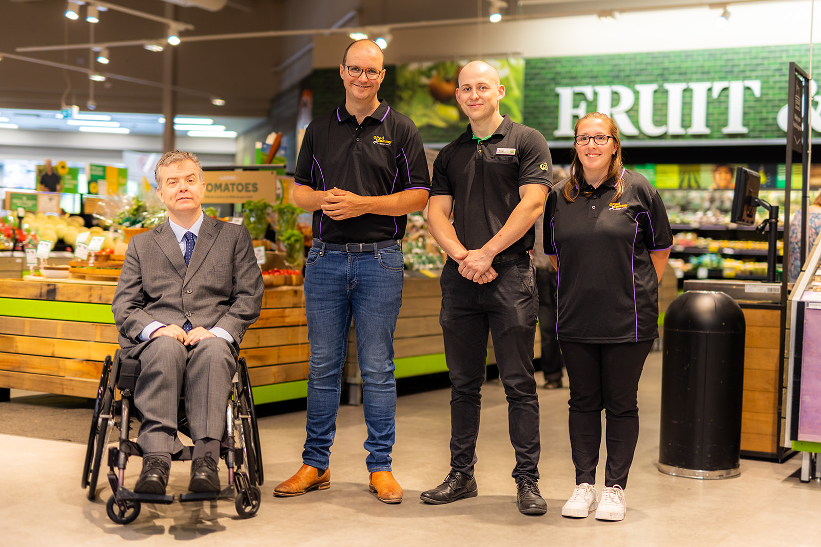 Photo of four people in a supermarket smiling at the camera. Once man is in a wheelchair. 