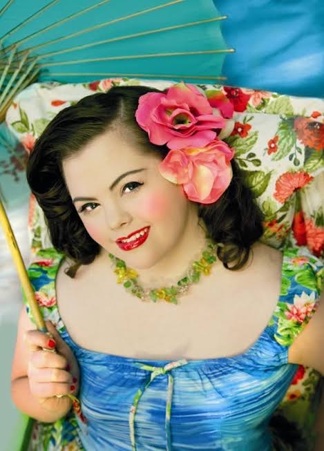 Image of Audrey wearing a blue dress and flower necklace holding an umbrella. She is wearing bright res lipstick, her hair is in curls and she has beautiful large pink flowers behind her ear. 