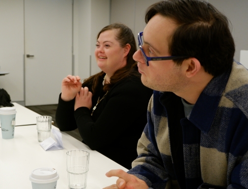close up of a male and female side on sitting at a table. The female in the background is smiling. The man in the foreground is wearing glasses. 