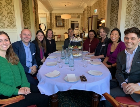 A group of people sitting around a table with a white table cloth having afternoon tea. They are smiling at the camera 