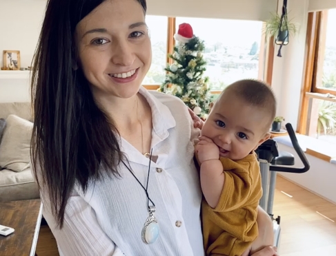 Heidi is standing and holding her baby. She has long dark hair and wearing a white shirt. She is smiling at the camera. 