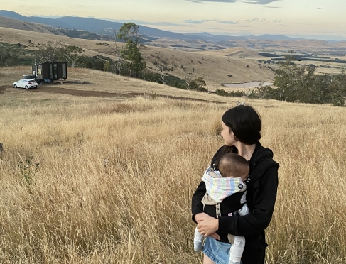 Heidi holding her baby looking into the distance. Heidi is standing in a field with mountains in the background 