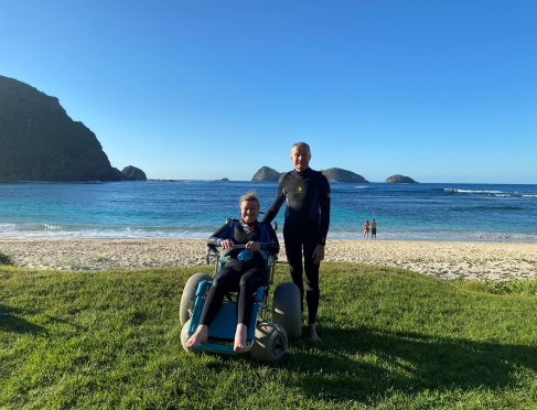 Image of Jane with another person. Jane is sitting in a wheelchair and the man is standing. They are on grass with white sand and blue water behind them. There are mountains behind the water. Both people are smiling. The sky is blue. 