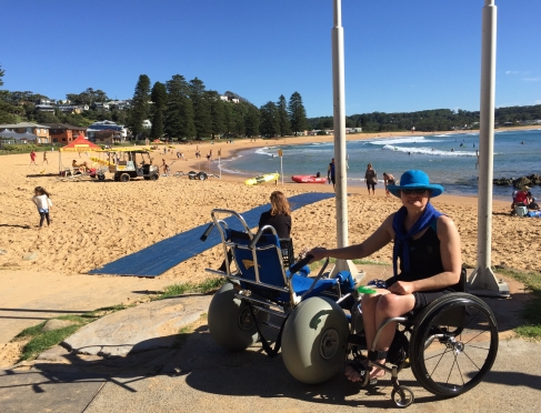 Image of Jane about to trasnfer into a beach wheelchair. She is wearing a blue hat. She is by the beach. It's a beautiful sunny day with the ocean and trees in the background.