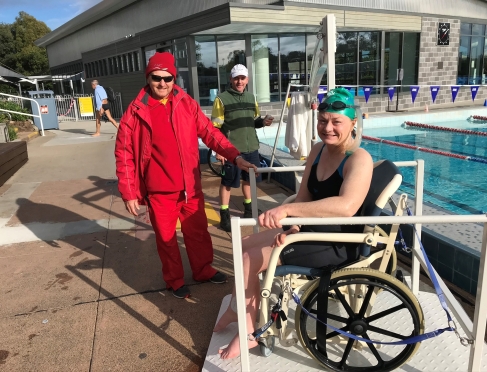 Image of Jane beside a pool. She is wearing a green cap and has goggles on. A man wearing red is standing with her. 