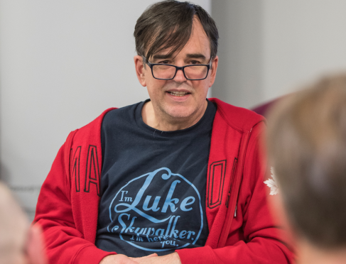 Image of Tim. He is wearing glasses, a red zip up hoodie and blue tshirt. He has his hands together on his lap and is speaking with a group of people