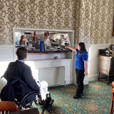 A trainee pointing to the kitchen. Commissioner Gauntlett is looking at the kitchen. He is sitting in a wheelchair