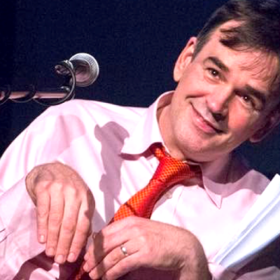 Image of Tim wearing a light shirt and red tie. He is in front of a microphone and looks as though he is performing. He is smiling and is making paw movements with his hands
