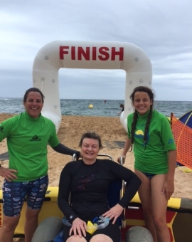 Image of Jane in a beach wheelchair with two young women either side of her in green. They are all smiling at the camera. There is a big finish line sign in the background on the beach.