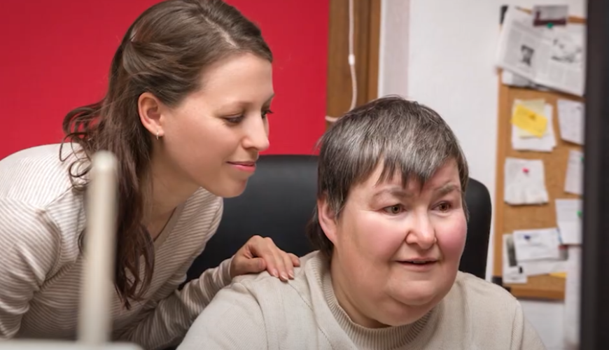 Image of a person in front of a computer. Next to this person is a female with a smile on her face. Her hand is on the person in front of the computer's shoulder. The image portrays a woman assisting a person using a computer.