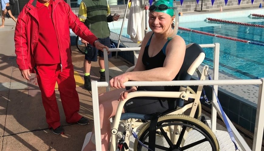 Image of Jane at the pool. She is wearing a swimming cap and goggles on her head. She is smiling at the camera. Jane is sitting in a wheelchair beside the pool. There is a man standing next to her smiling and wearing red 