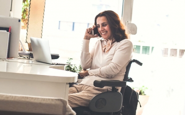 A woman in a wheelchair is smiling while she is on a phone call. She is working from home on her laptop.