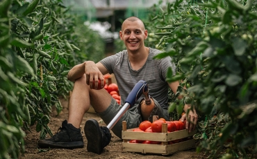 A young man with a prosthetic leg is sitting on the ground between rows of tomato plants. He is picking tomatoes and smiling