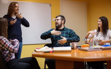A group of men and women are in a staff meeting. They are all communicating  together through sign language.