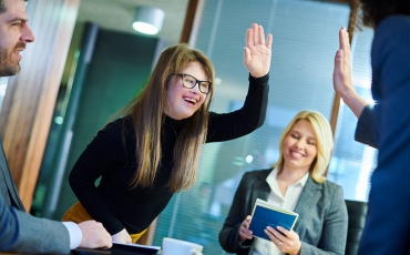 Two colleagues are raising hands for a high five. One is partially out of frame and the other is a young woman with long blonde hair and Down Syndrome.  A male colleague is smiling and  a woman is also smiling and looking down at a book.