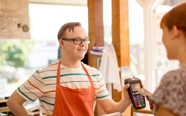 A young man with Down Syndrome is holding up an EFTPOS machine to a customer. He is working in a café. 
