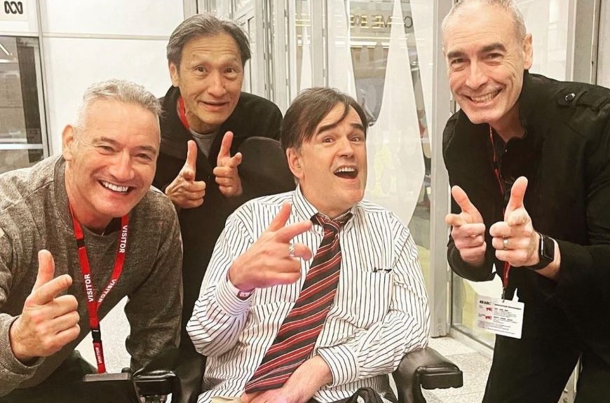 Image of Tim wearing a suit and tie and pointing his finger. He is with members of the wiggles 